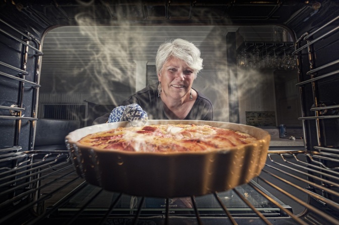 Eine lächelnde Frau mit grauen Haaren nimmt einen dampfenden Apfelkuchen aus dem Ofen.