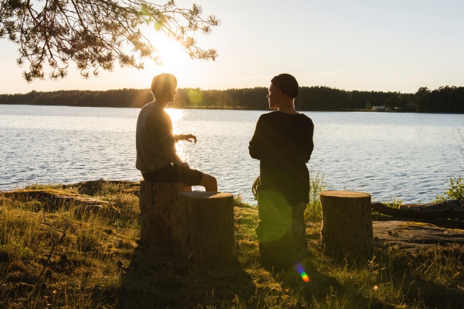 Ein Mann und eine Frau sitzen im Freien an einem See. Sie wurden im Gegenlicht fotografiert und sind nur als Silhouetten zu sehen.