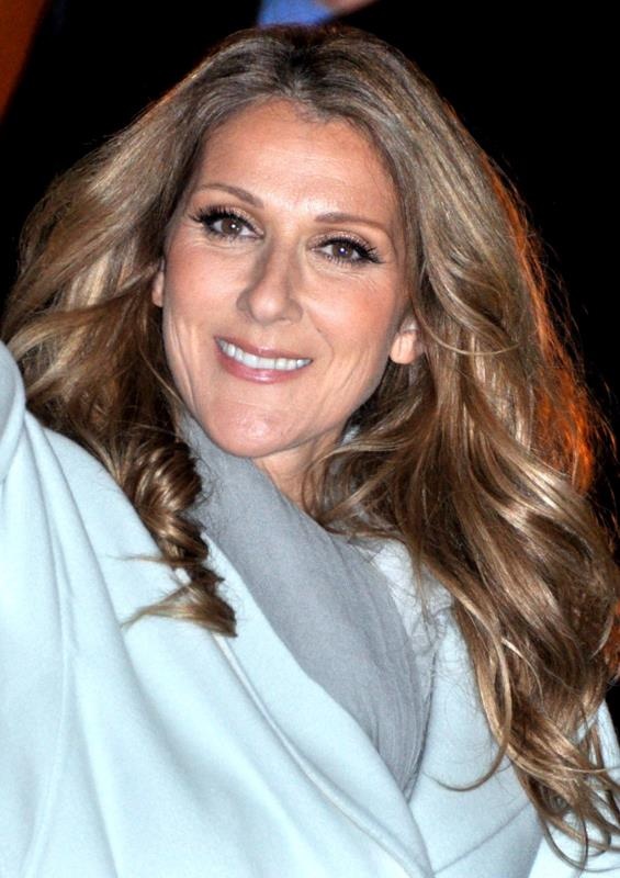 Céline Dion mit gewellten Haaren in heller Kleidung. Sie lächelt und winkt.