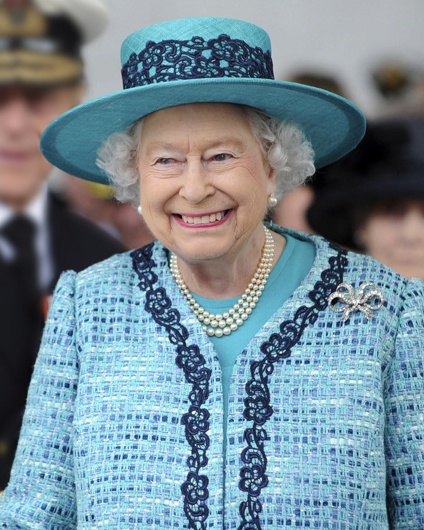 Queen Elisabeth die Zweite in einem Kostüm mit dazu passendem Hut. Sie lächelt breit.