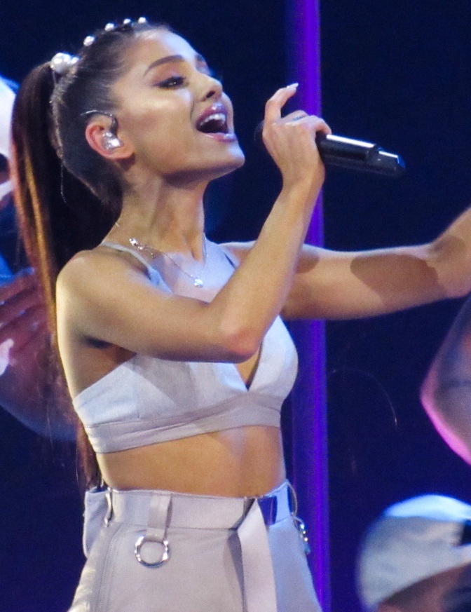 Ariana Grande in einem weißen Bühnen-Outfit und mit Pferdeschwanz. Sie singt in ein Mikrophon.