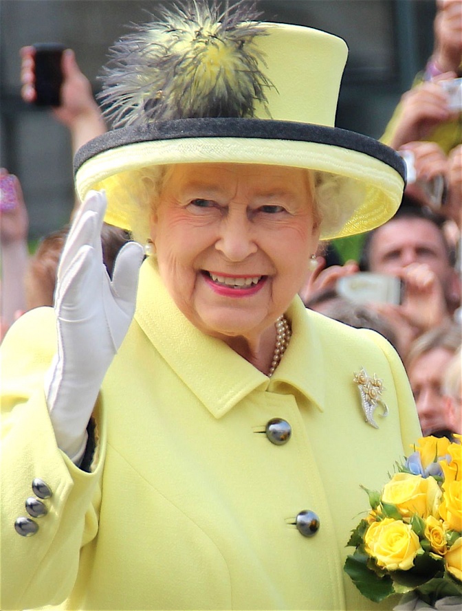 Queen Elisabeth in einem hellgelben Kostüm mit passendem Hut. Sie lächelt und hebt winkend die Hand. In der anderen Hand hält sie einen Blumenstrauß.