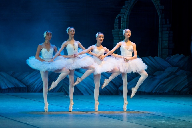 4 Tänzerinnen in weißen Tutus tanzen und haben sich beieinander eingehakt. Im Hintergrund sieht man ein Bühnenbild.