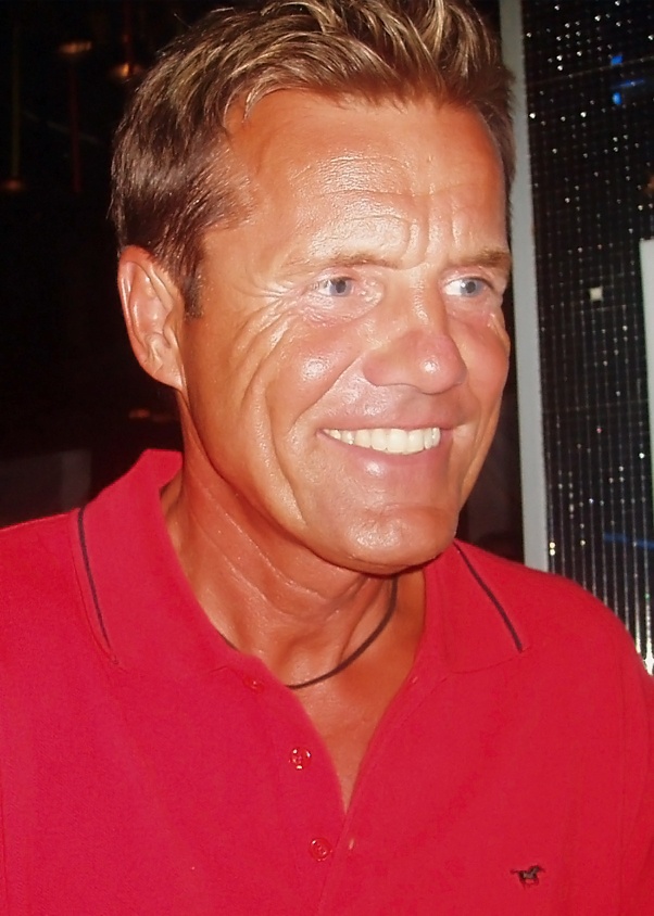 Dieter Bohlen mit gebräunter Haut und kurzen blonden Haaren. Er trägt ein rotes Poloshirt und lächelt.