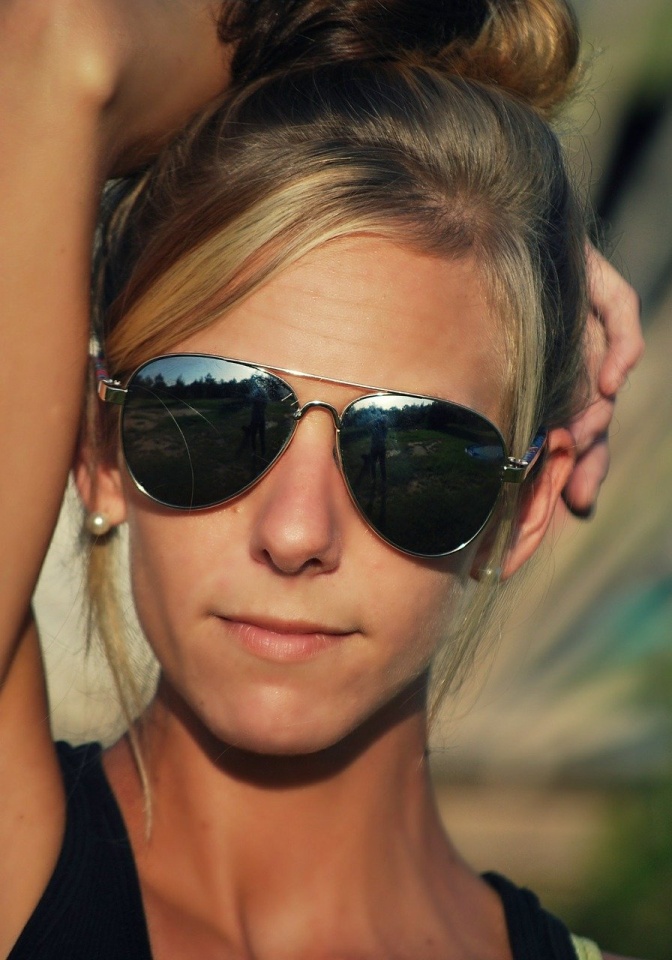Eine junge blonde Frau trägt eine Sonnenbrille in Pilotenform. Sie legt einen Arm über ihren Kopf und schaut in die Kamera.