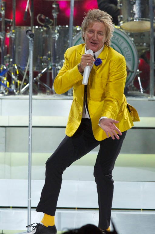 Rod Stewart auf der Bühne in einem gelben Sakko und mit blonden Haaren. Er singt in ein Mikro und tanzt.