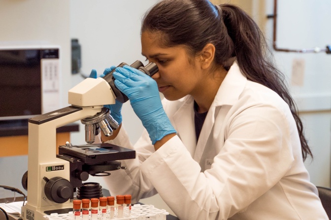 Eine Wissenschaftlerin mit dunkler Haut und einem schwarzen Pferdeschwanz sitzt in einem weißen Kittel an einem Mikroskop.