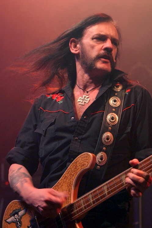 Lemmy Kilmister spielt auf der Bühne Gitarre. Die Bühne ist in Rot ausgeleuchtet. Er hat wehende, lange, braune Haare.