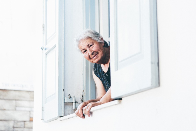 Eine alte Frau mit weißen Haaren schaut lächelnd aus einem geöffneten Fenster.