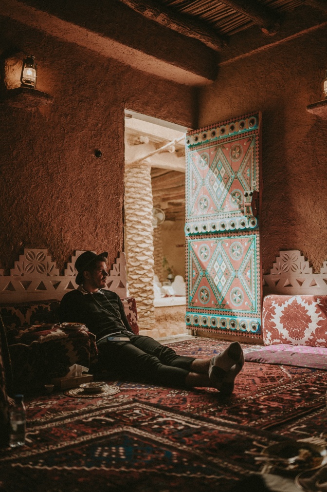 Ein Mann sitzt bei geöffneter Tür auf einem Teppich auf dem Boden eines Raumes. Er hat einen Hut auf und lehnt sich an die Wand.