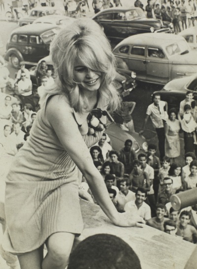 Brigitte Bardot als junge Frau. Sie lehnt sich lächelnd auf eine Brüstung. Im Hinterrund sieht man viele Menschen.