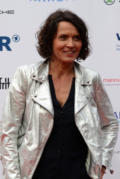 Ulrike Folkerts mit kinnlangen, gewellten Haaren und in glänzender Lederjacke vor einer Logowand