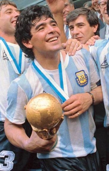 Diego Maradona in einem gestreiften Trikot. Er trägt eine Medaille um den Hals und hält einen Pokal in der Hand. Er steht zwischen seinen Teamkollegen.