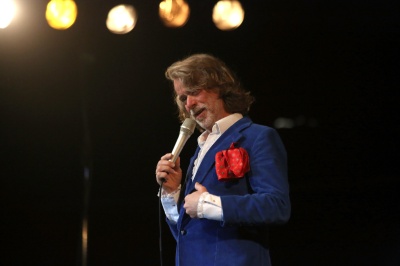 Helge Schneider spricht auf der Bühne in ein Mikrofon. Er hat schulterlange Haare und trägt einen Vollbart.