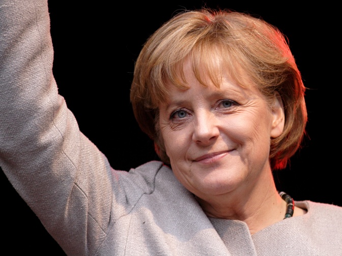 Angela Merkel in einem beigefarbenen Sakko. Sie winkt.
