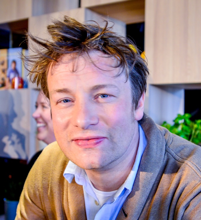 Jamie Oliver mit strubbeligen blonden Haaren. Er schaut direkt in die Kamera und lächelt leicht.