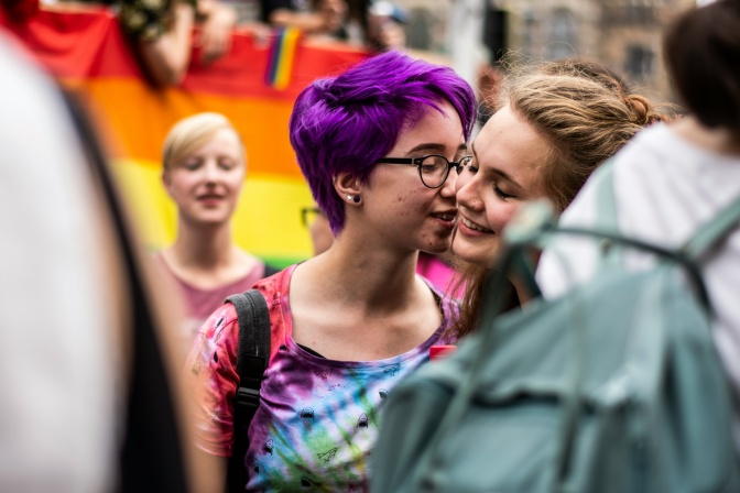 2 Frauen bei einer Pride Parade. Eine der beiden küsst die andere auf die Wange.