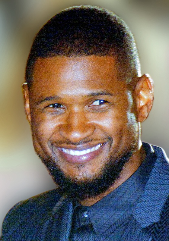 Der Sänger Usher lächelt ins Bild. Er ist dunkelhäutig und hat seine Haare sehr kurz rasiert. Er trägt einen Anzug mit Fischgrätmuster und eine dazu passende Krawatte.