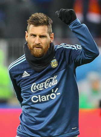 Lionel Messi in einem blauen, langärmligen Trikot. Er hebt den rechten Arm.