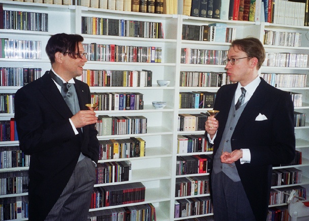 2 Männer im Morning Dress: Sie stehen vor einem Bücherregal, halten ein Getränk in der Hand und unterhalten sich.