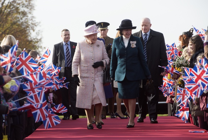 Queen Elisabeth und ihre Begleiterin im Kostüm. Sie laufen über einen roten Teppich. Rechts und links von ihnen stehen Blumengestecke.