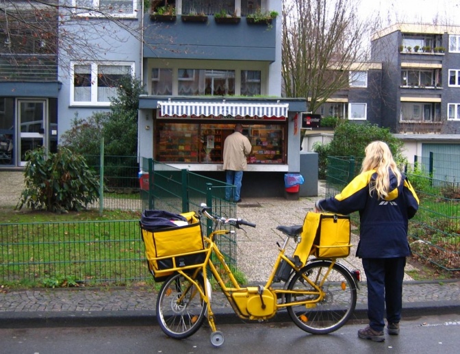Eine Brief-Trägerin der Deutschen Post ist mit ihrem Post-Fahrrad unterwegs. Sie ist von hinten zu sehen und läuft auf ein Haus zu.