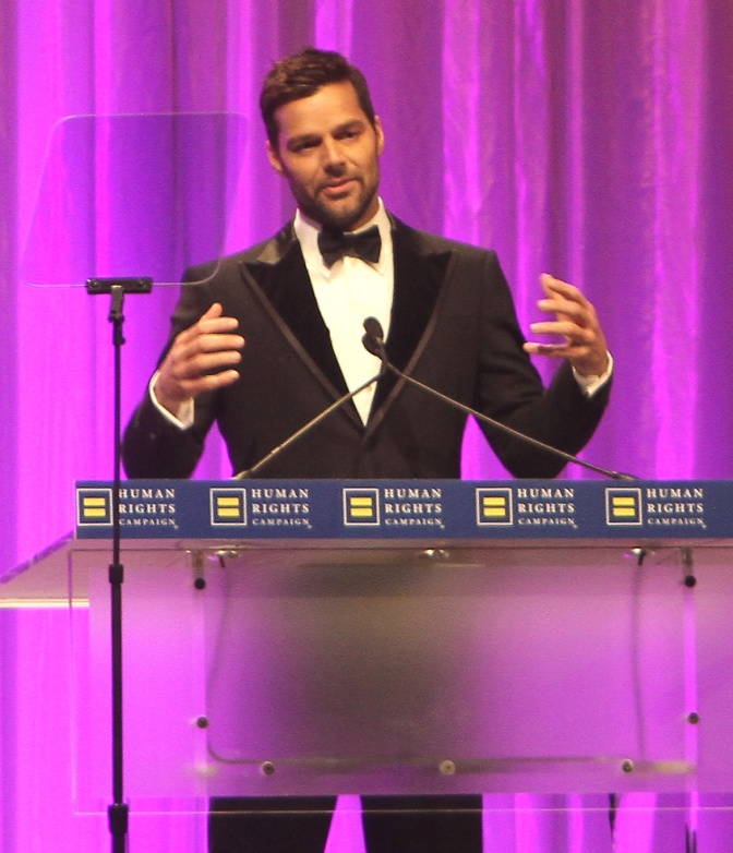 Ricky Martin steht vor einem grellen Vorhang an einem Rednerpult und spricht in ein Mikrophon. Er gestikuliert mit den Händen.