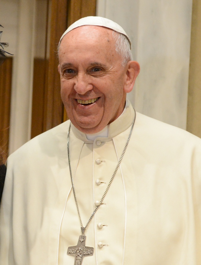 Papst Franziskus mit Papstkappe und weißem Talar. Er lächelt. Um seinen Hals hängt eine Kette mit einem großen Kreuz.
