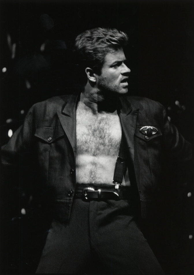 Ein altes schwarz-weiß-Bild von George Michael mit freiem Oberkörper. Er breitet die Arme aus und schaut zur Seite.