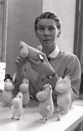 Ein schwarz-weiß-Bild von Tove Jansson. Auf dem Tisch vor ihr stehen Mumin-Figuren, eine davon hält er in der Hand.