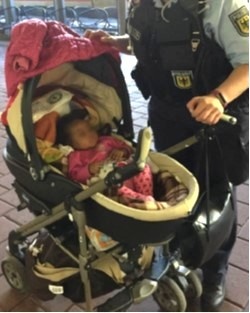 Ein 5 Monate altes, dunkelhäutiges Baby im Kinderwagen. Eine Polizistin in Uniform steht neben dem Kinderwagen.