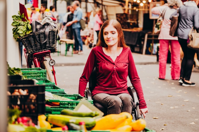 Eine Frau im Rollstuhl unterwegs in Berlin. Sie steht an einem Gemüsestand.