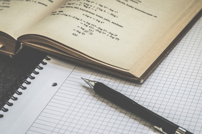 Ein aufgeschlagenes Buch mit Matheaufgaben, daneben ein karierter Schreibblick und ein Kugelschreiber