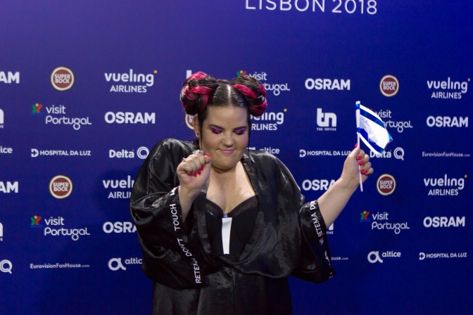 Netta hält die Flagge Israels mit geschlossenen Augen in der Hand. Sie steht vor einer Wand mit Logos von Fenrseh-Anstalten.