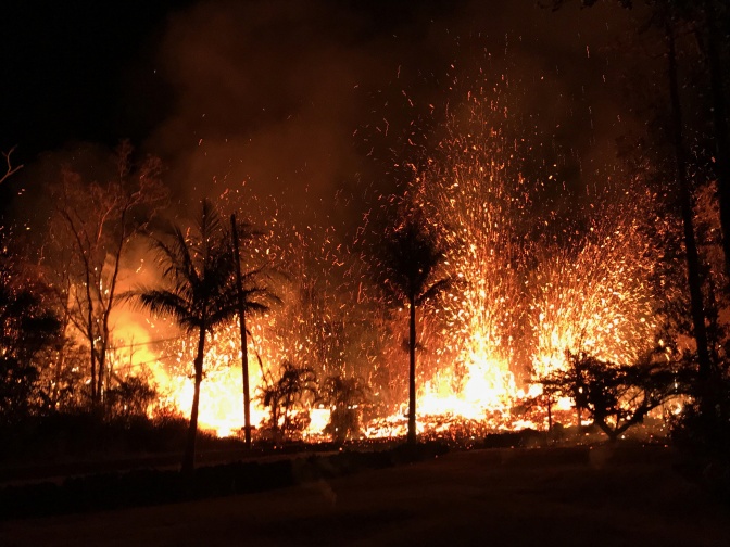 Ein nächtlicher Ausbruch des Vulkans Kīlauea. Im Vordergrund sieht man Palmen.