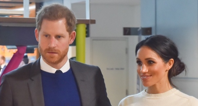 Prinz Harry und Herzogin Meghan stehen nebeneinander. Er trägt Hemd und Pullover, sie ein helles Oberteil.