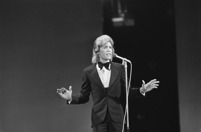 Ein schwarz-weiß-Foto von Jürgen Marcus in Anzug und Fliege vor einem Mikrophon. Er hat eine Vokuhila-Frisur.