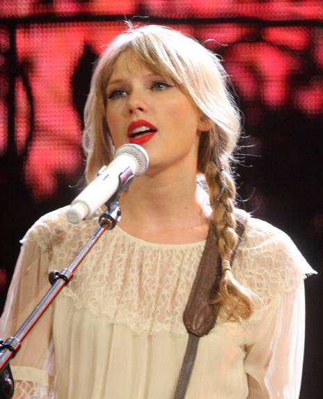 Taylor Swift mit rotem Lippenstift und  in einer Spitzenbluse mit geflochtenen Zöpfen. Sie singt in ein Mkrophon.