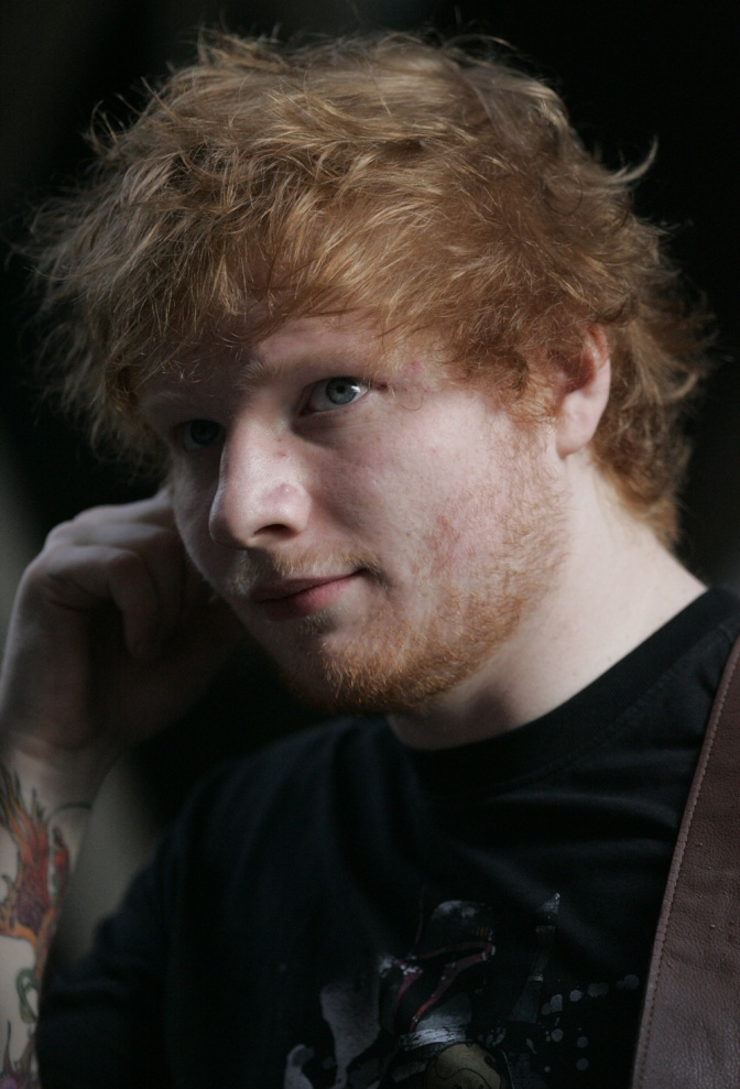 Ed Sheeran in einem dunklen T-Shirt vor dunklem Hintergrund. Man sieht mehrere Tattoos am Arm.