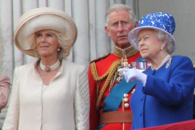 Prinz Charles trägt Uniform, die beiden Damen farbige Kostüme mit passenden Hüten. Sie stehen auf einem Balkon. Die Queen winkt.