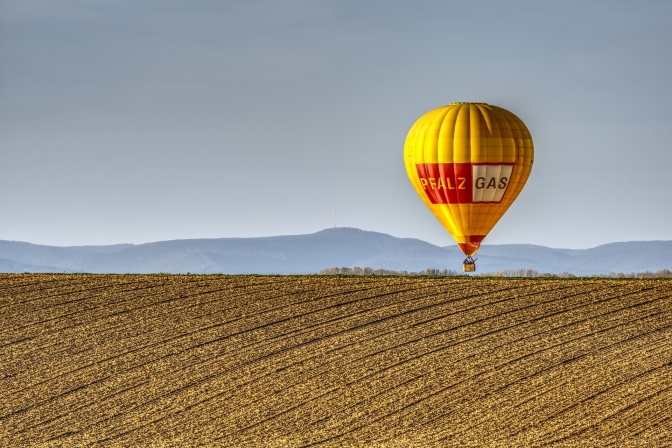 Ein Heißluftballon fliegt über ein abgeerntetes Feld, im Hintergrund ein Bergzug