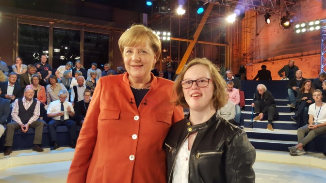 Angela Merkel und Natalie Dedreux stehen nebeneinander. Beide schauen in die Kamera und lächeln. Im Hintergrund sitzen Zuschauer und Zuschauerinnen auf Stuhlreihen.