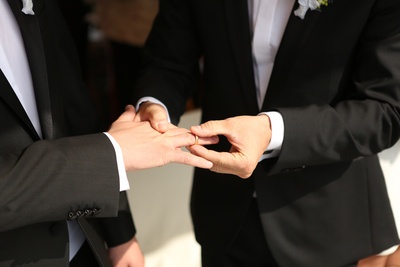 Zwei Männer in schwarzen Anzügen stecken sich gegenseitig Eheringe an.