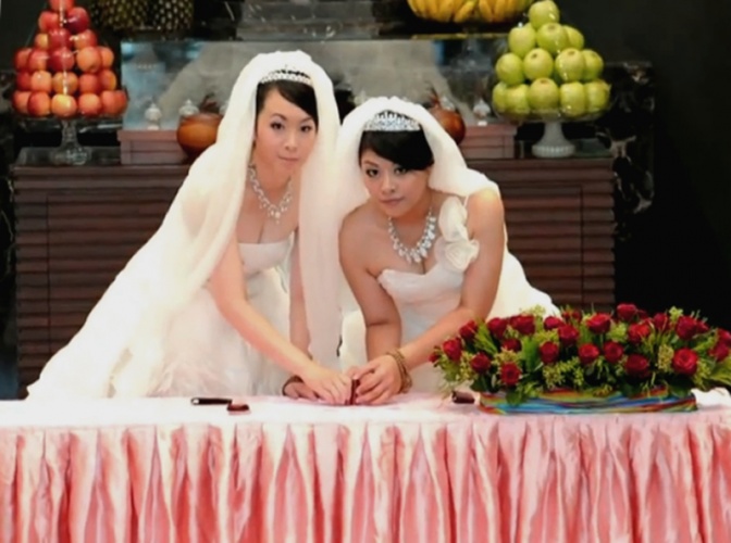 2 asiatische Bräute in weißen Kleidern und mit Schleier tauschen Eheringe. Neben ihnen steht ein Gesteck mit roten Rosen.