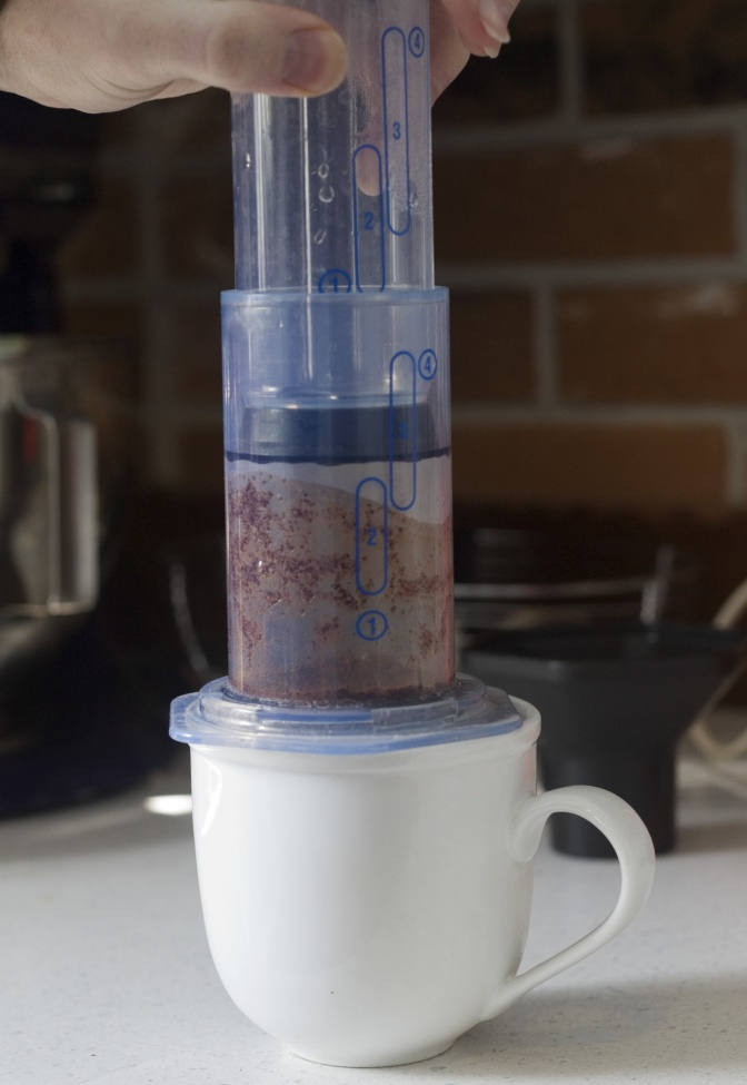 Eine Aeropress aus Plastik steht auf einer weißen Kaffeetasse. Im Inneren befinden sich gemahlener Kaffee und Wasser.