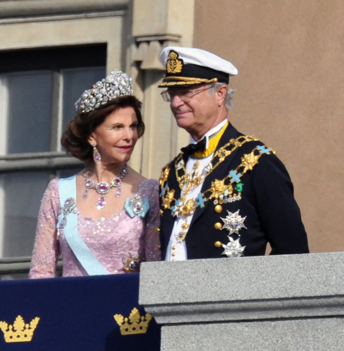 Das schwedische Königspaar steht auf dem Balkon. König Carl Gustaf trägt eine Uniform mit Mütze, Königin Silvia ein elegantes, langärmliges Kleid. Sie trägt ein Diadem und zahlreiche Juwelen. Er trägt Orden und große goldene Ketten.