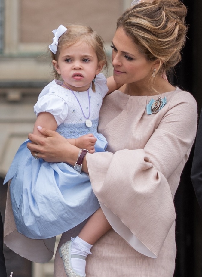Madeleine von Schweden trägt ein Kleid in hellrosa mit Trompetenärmeln. Sie trägt ihre Tochter auf dem Arm und spricht mit ihr.