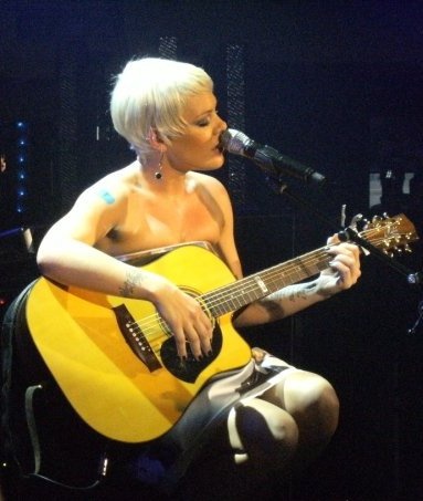 P!nk sitzt auf einem Hicker, spielt Gitarre und singt in ein Mikrophon. Ihre Haare sind kurz und blondiert.
