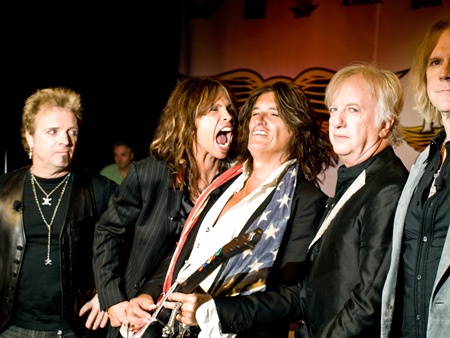 Die Mitglieder der Band Aerosmith stehen in schwarzen Lederjacken nebeneinander in einer Reihe. Steven Tyler reißt den Mund weit auf.