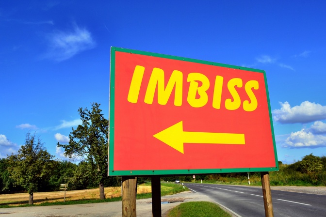 Ein Hinweisschild mit einem Pfeil und dem Wort Imbiss. Im Hintergrund Bäume und blauer Himmel.
